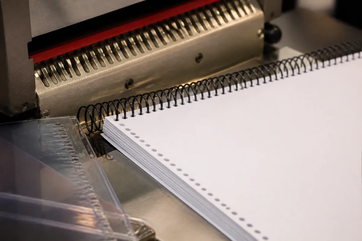 Gros plan sur une reliure spirale en cours de pose, avec des feuilles A4 perforées empilées et un dos de couverture transparent posé à côté